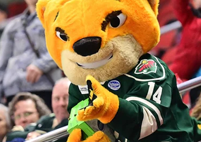 Cheerful mascot in a green hockey jersey, playfully pointing at the camera during a lively sports event.