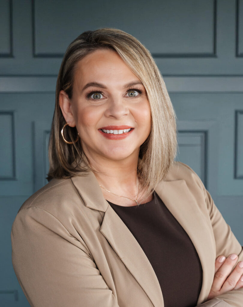 Professional woman smiling in a beige blazer with arms crossed against a blue paneled background.