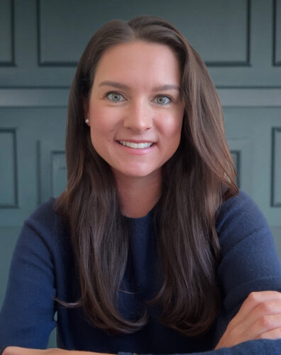 Smiling woman with long brown hair, wearing a navy sweater, against a paneled background.