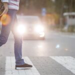 Person crossing street at crosswalk with car headlights shining. Carrying bag of groceries.